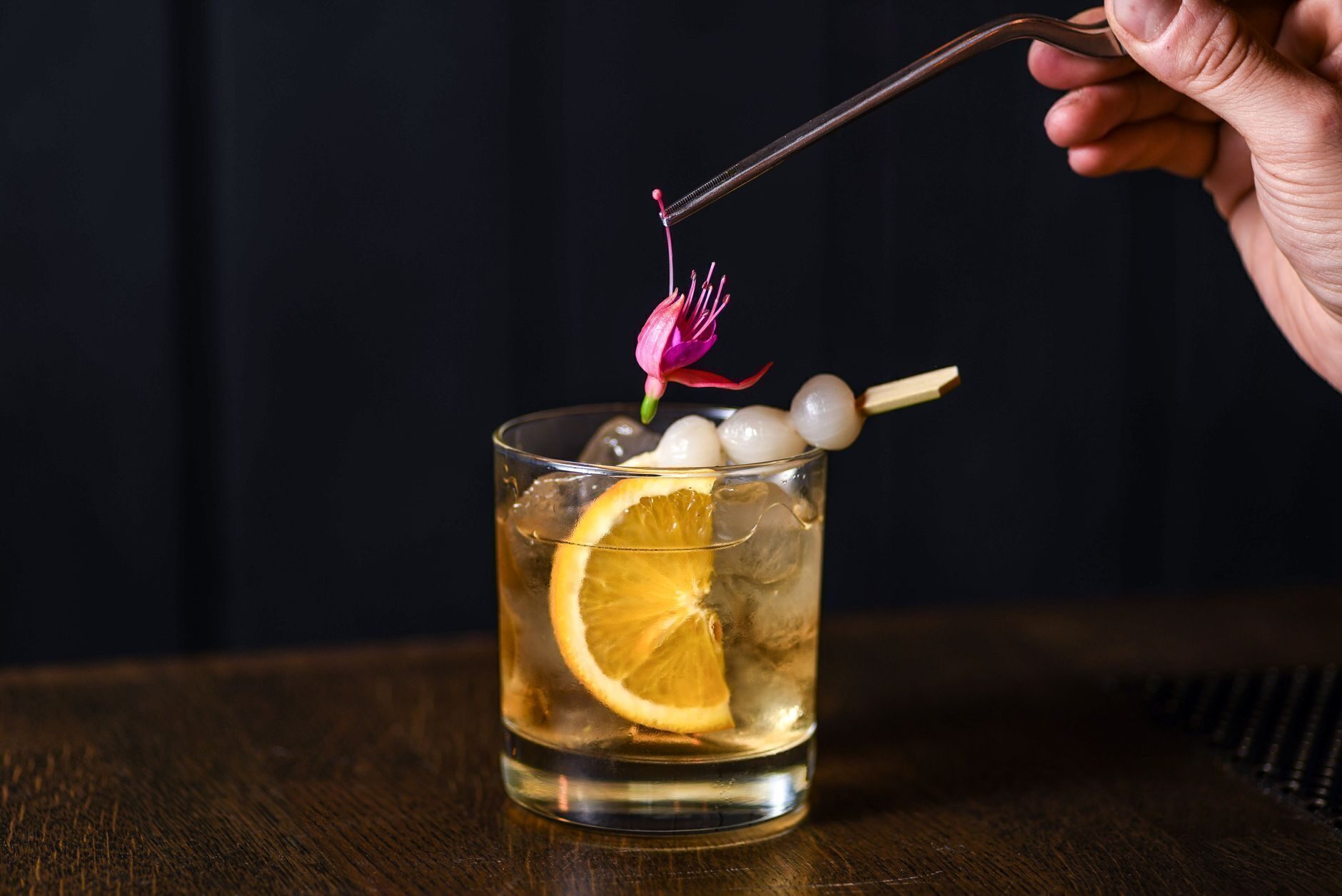 A smoked bourbon old fashioned garnished with an orange wheel and edible flower on dark wood